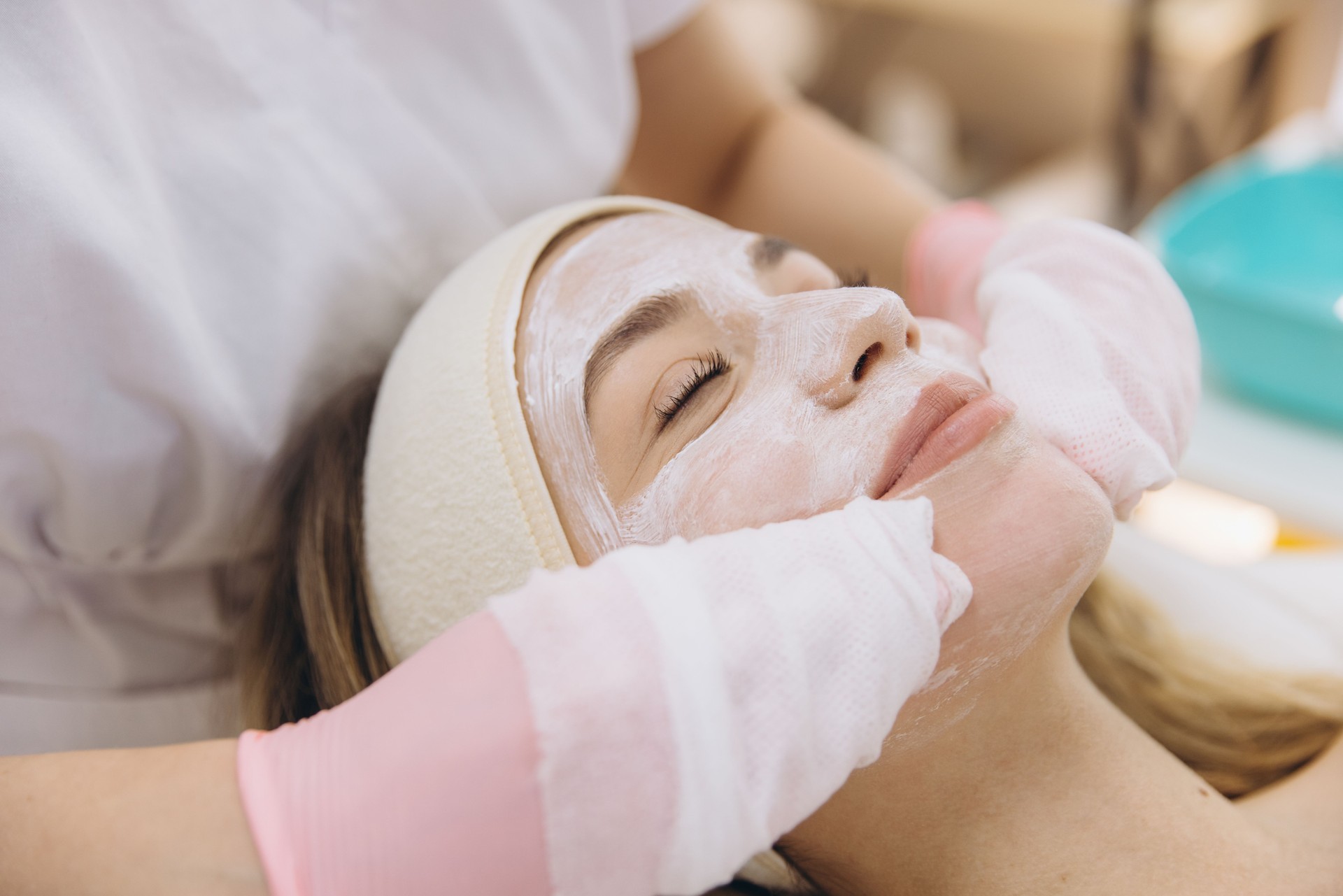 Beautician wearing gloves applying a white facial mask on the face of a young woman lying on a bed in a beauty salon